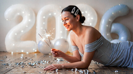A European-looking girl lies on the floor with balloons in the background. She holds sparklers and wears a silver dress and pearl hairpins.
