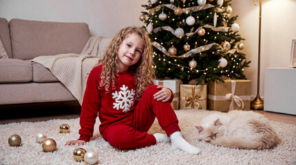 The girl is sitting on the floor on a fluffy white carpet. She is wearing red pajamas with an appliqu&eacute;. A cat is lying nearby.