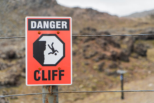 Close up of danger cliff sign. Bold red, black and white sign with graphic symbol of person falling off a cliff.  Background of bare rocky hills.  Horizontal landscape orientation.