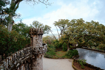 Medieval style stone tower and crenellated wall rising above lush green trees and garden path on Sintra hillside.