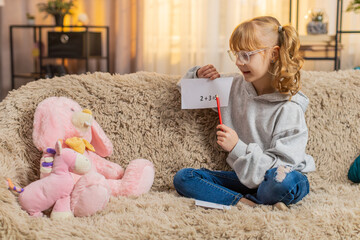 Teenager girl places toys plush figures in front at home sofa, seriously reciting repeats simple math arithmetic. Child kid occasionally giggles laughs amused by own mock authority, silly teacher role