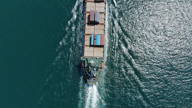 Top down aerial view of light loaded container ship sailing at sea channel. Camera look straight down, flying faster than cargo carrier, showing upper deck from aft to fore.