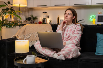 Middle-aged woman at night home sits with laptop thinks about task remote work focus calm. Caucasian girl on sofa at evening reflects plans idea drafts notes types research online session quiet mood.