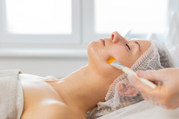 Fototapeta premium Caucasian young woman lies on couch while cosmetologist applies facial mask with brush on face in clinic. Relaxation skincare treatment gives smooth skin comfort wellness and calm mood during session.