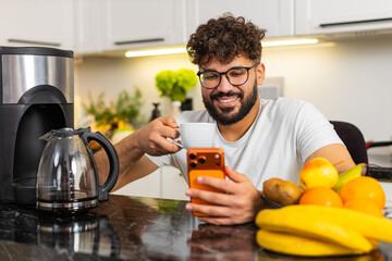 Arabian man drinks coffee in kitchen at home enjoying strong aroma craving caffeine for morning focus and routine. Young guy checks news on smartphone scrolling social media showing coffee addiction.