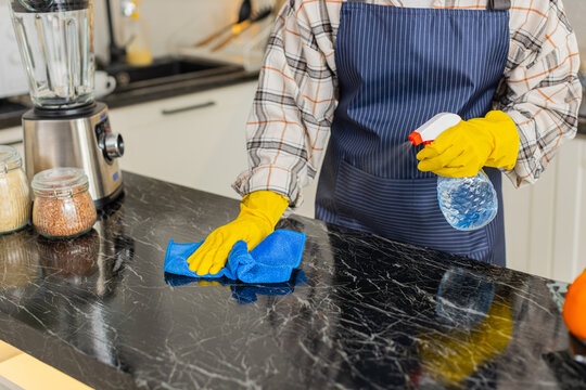 Woman wipes kitchen counter with disinfectant spray and cloth wearing yellow gloves for daily home hygiene. Girl completes quick cleanup keeping own apartment kitchen safe for meal prep indoors.