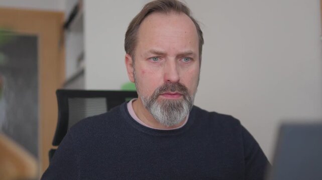 A handsome middle-aged man works on a laptop and drinks a cup of coffee in an apartment