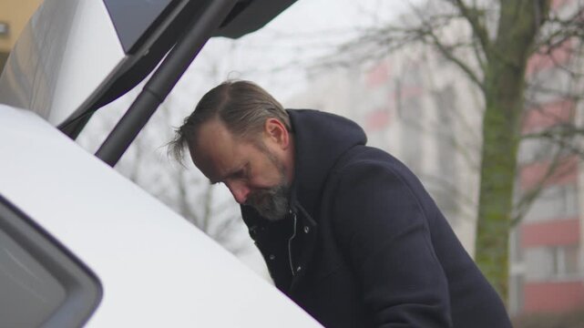 A handsome middle-aged man arranges the contents of the trunk of his car, presses a button which closes the trunk