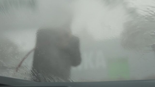 A man power washes his car with a power wash - view from the inside through the windshield sheeted with water