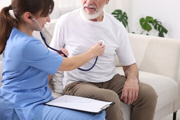 Obraz premium Care worker checking senior man's heartbeat with stethoscope on sofa at home, closeup