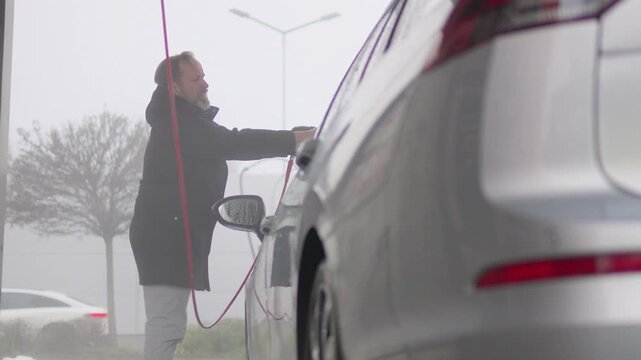 A middle-aged Caucasian man power washes the front of his car - low view from behind the car