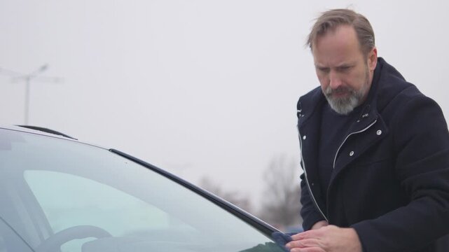 A handsome middle-aged man wipes a windshield with a cloth - view from the hood of the car
