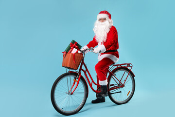 Santa Claus on bicycle with Christmas gifts in basket against light blue background