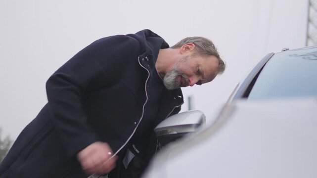 A handsome middle-aged man wipes the front window and side mirror of his car - view from the hood