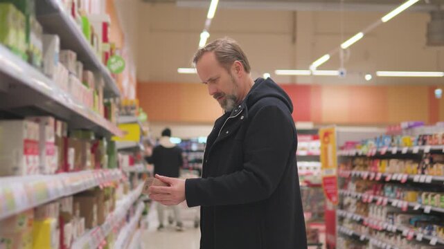 A handsome middle-aged man finds a product he likes, adds it into a basket and keeps looking in a supermarket