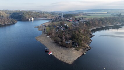 Aerial view shows a reservoir with a peninsula.  Residences sit on the land.  Trees populate the terrain.  A dam is visible.  The water reflects the sky.  The scene appears tranquil © rikajiMiAda