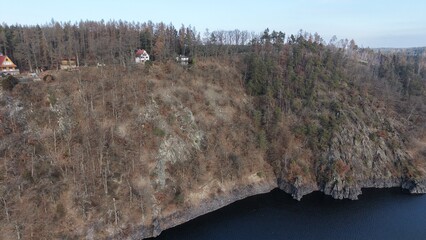 Aerial view shows a steep cliff face.  Trees cover the slope.  Residences sit atop the ridge.  A body of water is visible below.  The sky is clear.  The scene appears tranquil © rikajiMiAda