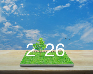 2026 white text with growing tree on green grass on open book on wooden table over blue sky with white clouds, Happy new year 2026 ecological cover concept