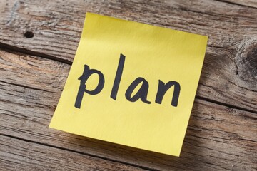 A vibrant yellow sticky note, featuring the word "plan" clearly handwritten in bold black ink, rests on a naturally textured, weathered wooden surface. This straightforward image evokes concepts of st