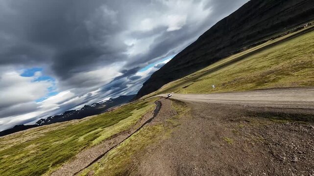 Adrenaline-pumping FPV chase over Iceland's dramatic landscape. The drone dives low over a small stream and follows a white van as it navigates a winding mountain pass under a vast, moody summer sky