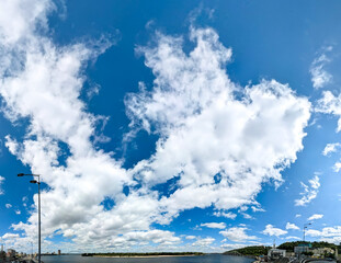 Beautiful blue sky background with cumulus clouds over the river and the city panorama. Large and small vapor structures are arranged in chain, flowing smoothly along the horizon. Wide website banner.