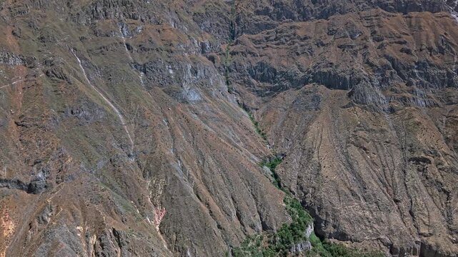 Drone approaches Colca Canyons barren sheer cliffs and rugged rock walls focusing on thin strip of vivid green vegetation. Water flowing from Cabanaconde brings life to otherwise unliveable landscape
