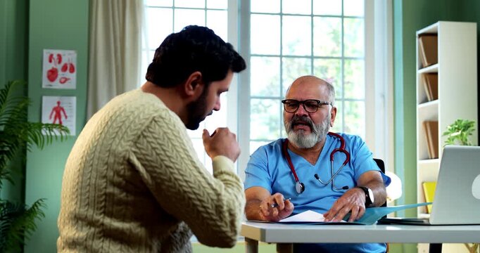 Indian male doctor discussing prescription with young male patient while writing medical advice as experienced physician explains treatment plan inside modern Indian clinic OPD healthcare visit
