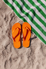 Top view of orange flip-flops on sandy beach next to green striped towel