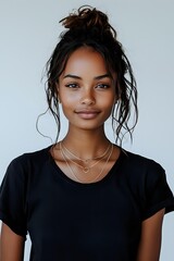 Young mixed-race woman with messy bun hairstyle wearing black t-shirt and layered gold necklaces smiling at camera against neutral background.