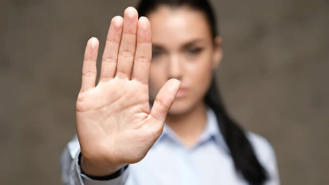 A woman makes a clear stop gesture with her hand while wearing a blue shirt, set against a neutral grey backdrop in a studio setting.