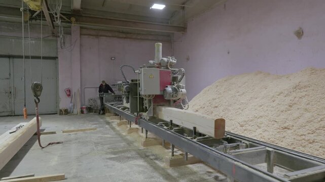 Long wooden beam being cut by industrial saw in a sawmill, with a large pile of sawdust beside it. Indoor workshop setting with visible tools and equipment
