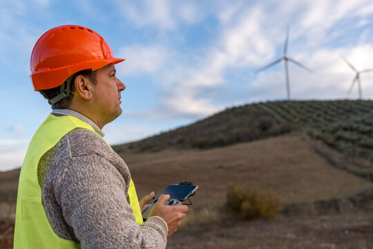 Worker in safety gear remotely controlling a drone against a backdrop of wind turbines on a hill, symbolizing modern energy