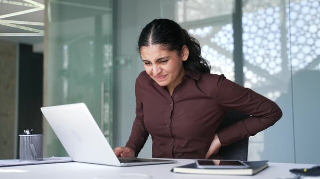Tired businesswoman suffering from back pain while working on laptop sitting at workplace in a business office. Upset young female employee holds her hand on her lower back, massaging her sore muscles
