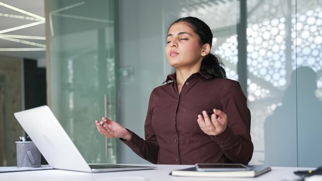 Young businesswoman meditates with her eyes closed sitting at workplace in business office. Woman employee took break from work on laptop. Calm woman rests, relaxes, feels peace of mind