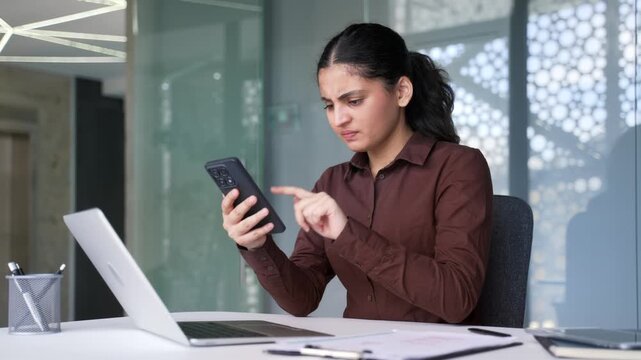 Angry young businesswoman having issues with using mobile phone app at workplace in business office. Confused female feeling annoyed with low battery, problems with smartphone or unstable connection.