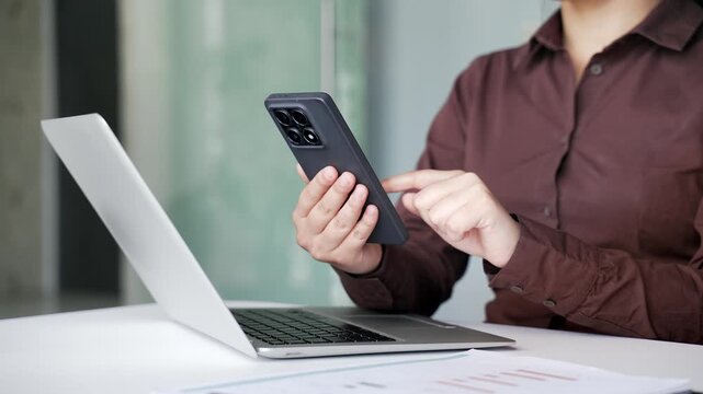 Close up of businesswoman's hands using mobile phone while sitting at desk with laptop at workplace in business office. Female employee reading message, chatting online with a client or browsing web