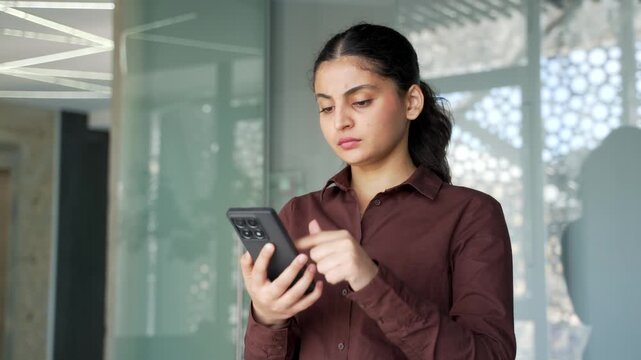Serious young businesswoman is using mobile phone sitting at a workplace in a modern business office. Thoughtful female employee reading a message, chatting online with a client or browsing the web. 