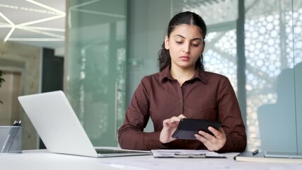 Focused businesswoman sitting at desk using calculator, writing notes, and working on laptop. Woman financial planning, accounting tasks, budgeting, data analysis, and concentrated office workflow