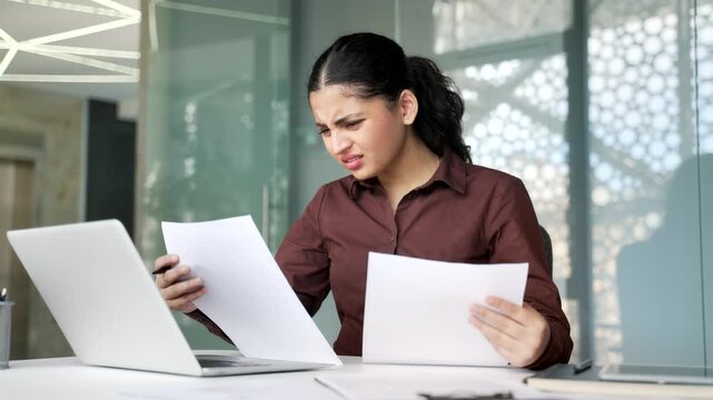 Confused young businesswoman having difficulty with paper work sitting at workplace in business office. Shocked female employee looks at documents and cannot understand problem and looking at camera