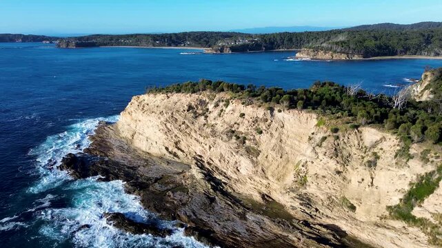 Drone aerial landscape with ocean waves crashing on rocky headland cliffs rugged terrain covered bushland trees overlooking Rosedale suburban town in Malua Bay South Coast Australia nature tourism