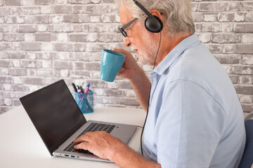Senior man working at desk,  using laptop in home office while drinking a coffee. Concept of active aging, retirement management, remote work, technology use by modern older adults