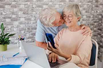 Affectionate older couple, the husband kissing his wife while she checks household bills on the computer and uses her cell phone. Two modern retirees using new technology.