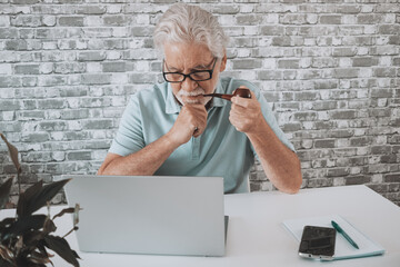 Portrait of old senior bearded man smoking pipe at home sitting at table with laptop. Elderly male surfing the net with computer