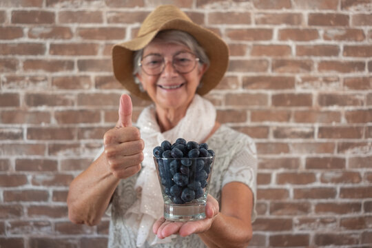 Smiling senior woman holding a glass of fresh blueberries and showing a thumbs-up gesture. Healthy eating concept, positive aging, nutrition, and wellbeing, with a brick wall background - Powered by Adobe