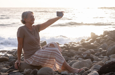 Portrait of relaxed senior woman sitting at the beach at sunset smiling looking at smartphone taking a selfie - elderly lady enjoying vacation and freedom