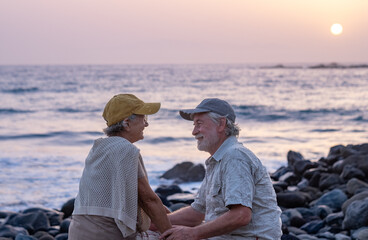 Bonding senior couple hands in hands while sitting on a rocky beach face the sea at sunset light expressing love and care. Retirement lifestyle, vacation, freedom