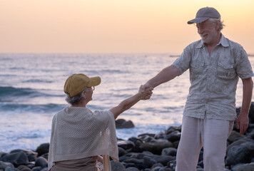Bonding senior couple hand in hand while sitting on a rocky beach face the sea at sunset light expressing love and affection. Retirement lifestyle, vacation, freedom