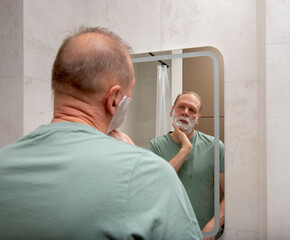 A mature man checks his face while applying shaving foam and looking at his reflection in a bathroom