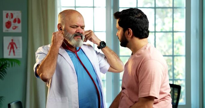 Indian male doctor treating patient during routine health checkup as experienced physician examines young man with stethoscope in modern Indian clinic OPD healthcare consultation visit