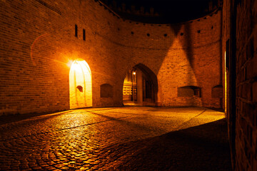 Warm lighting at curved brick walls of historic fortress at night, casting dramatic shadows across cobblestones. Arched doorways and textured masonry evoke timeless atmosphere